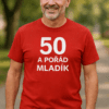 Muž v červeném tričku s nápisem 50 a pořád mladík, outdoorové prostředí, sportovní móda, aktivní stárnutí.