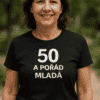 Mladá žena v tričku s nápisem "50 a pořád mladá" vystupuje v přírodním prostředí, ukazuje aktivní a zdravý životní styl po padesátce.