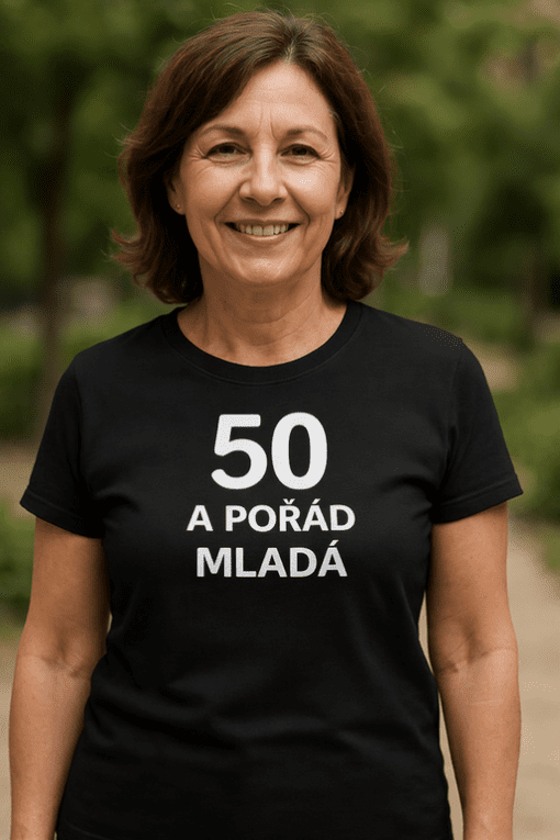 Mladá žena v tričku s nápisem "50 a pořád mladá" vystupuje v přírodním prostředí, ukazuje aktivní a zdravý životní styl po padesátce.