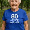 Starší žena v modrém tričku s nápisem „80 a pořád jako lux“ při venkovní aktivitě, zdravý životní styl, seniorská hygiena a energie.