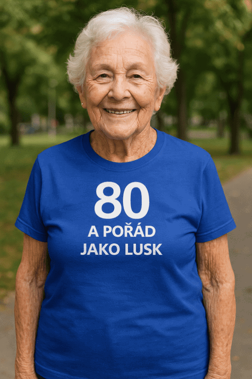 Starší žena v modrém tričku s nápisem „80 a pořád jako lux“ při venkovní aktivitě, zdravý životní styl, seniorská hygiena a energie.