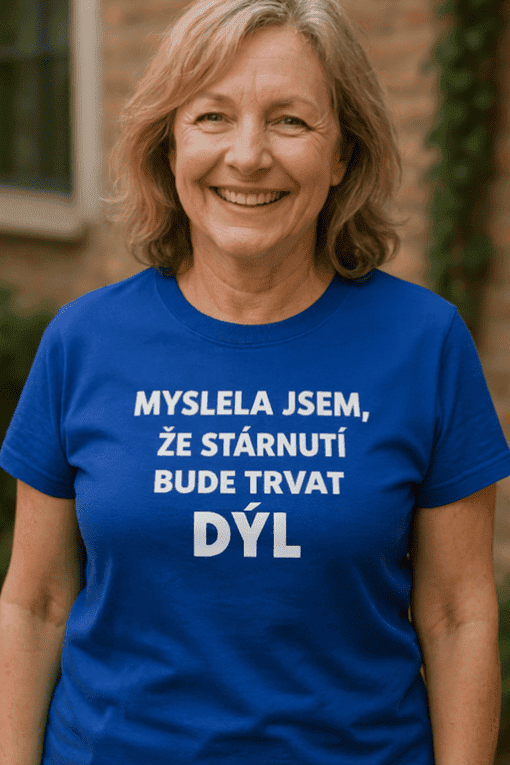 Žena v modré tričku s nápisem o stárnutí, přátelský výraz, přírodní pozadí.