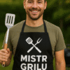 Pohled na muže v černém grilovacím tričku s nápisem MISTR GRILU a kuchyňským náčiním, ideální pro grilování a outdoorové akce.