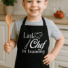 Vášnivý malý kuchař v černé zástěře s nápisem Little Chef in training, připraven na vaření.