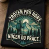 Tričko s horalským motivem v adventure badge stylu a textem Zrozen pro hory. Nucen do práce., s horami, sluncem a borovicemi v tyrkysovo-krémové potisku.